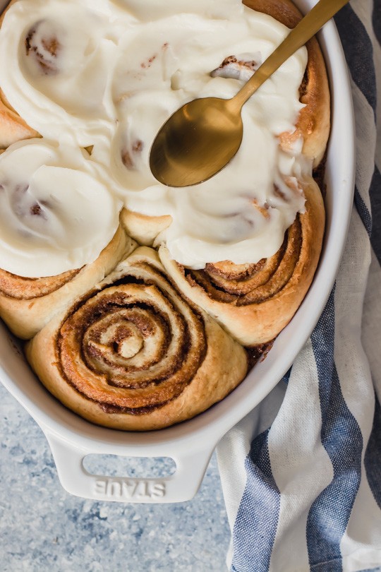 A pan of perfectly swirled cinnamon rolls with fluffy white frosting being spread over them.