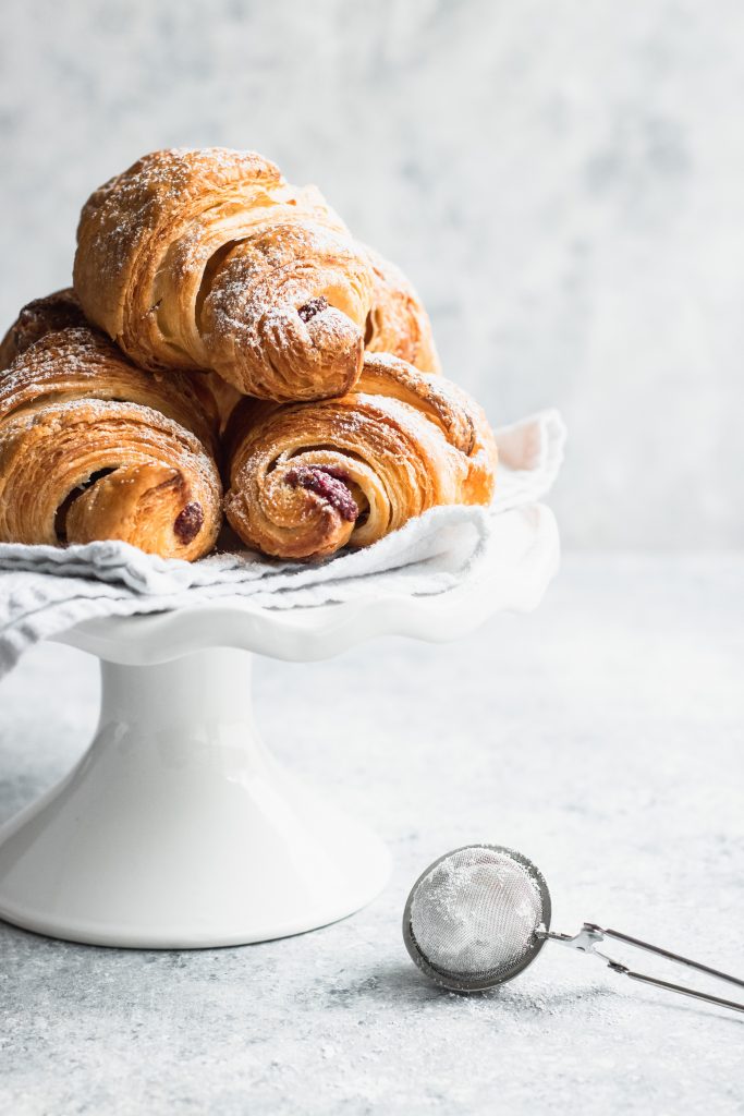 Piled on a white plate, these flaky croissants are filled with Chocolate and Raspberry Filling and dusted with powdered sugar.