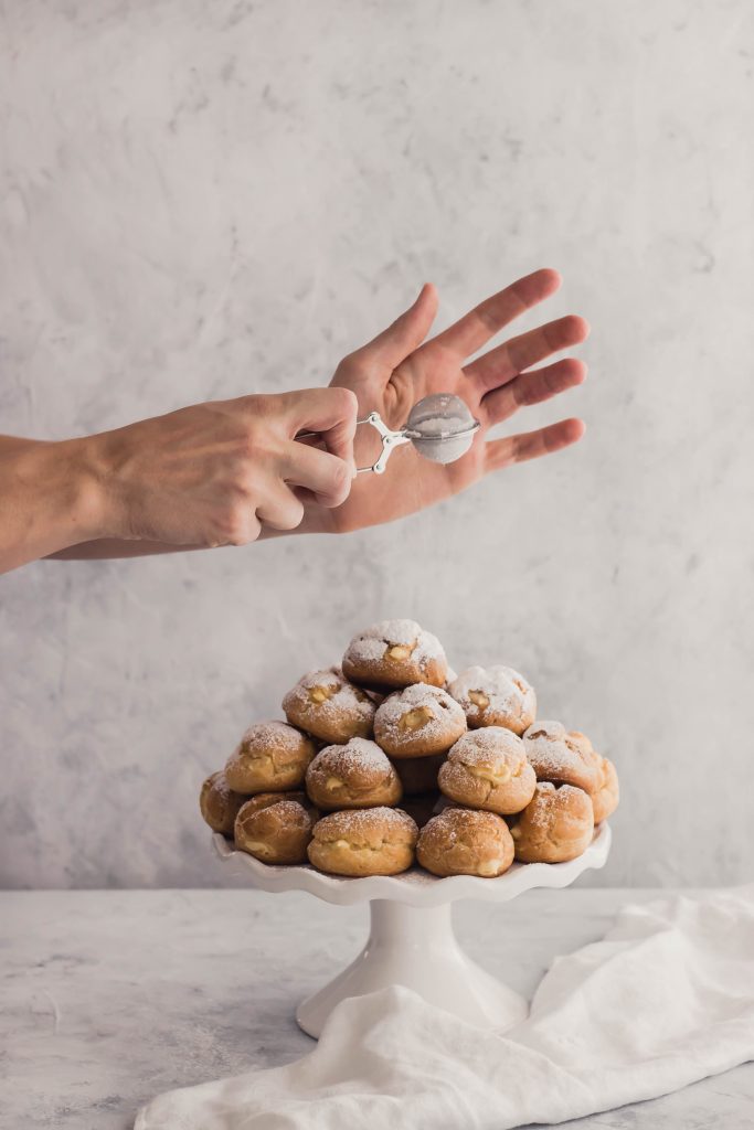 Maple cream puffs on a pedestal plate being dusted with powdered sugar