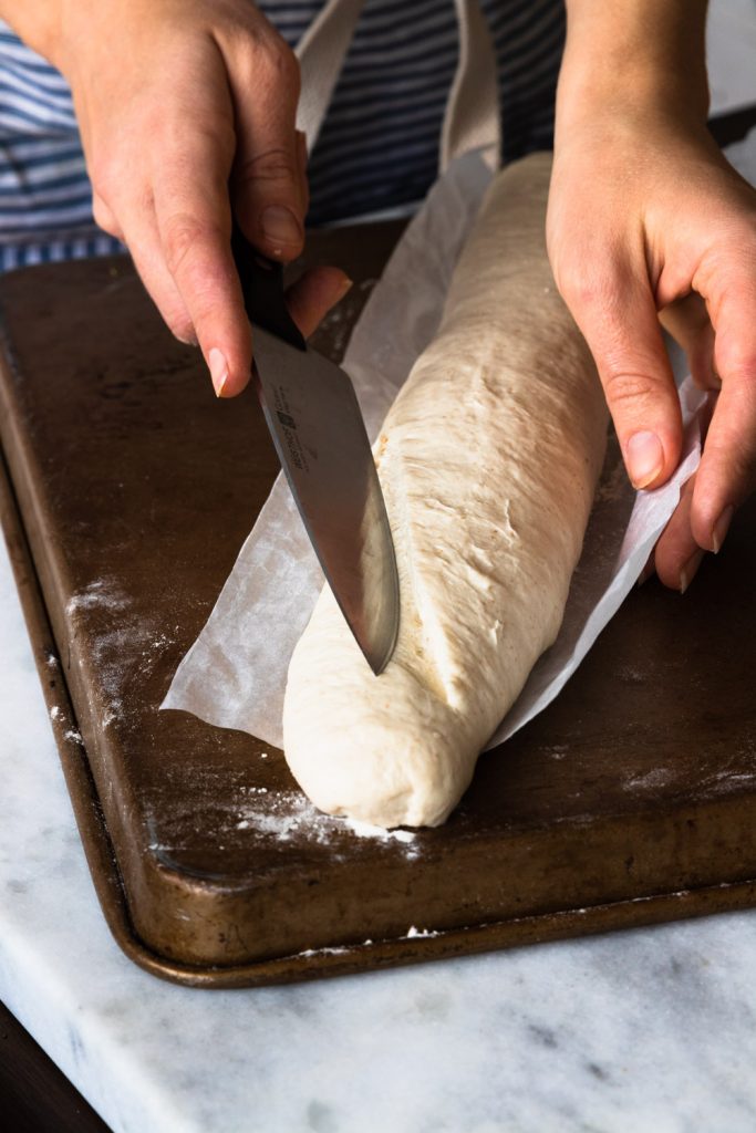 A baguette being slashed with a sharp chef's knife.