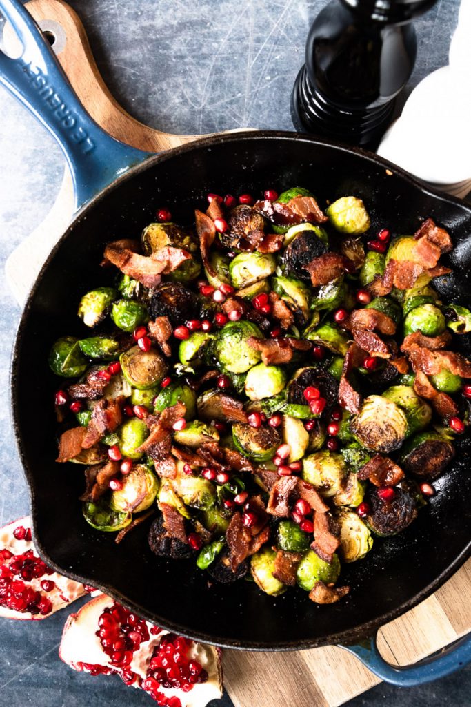 A pan of seared brussels sprouts and bacon, topped with pomegranate molasses and fresh pomegranate arils.