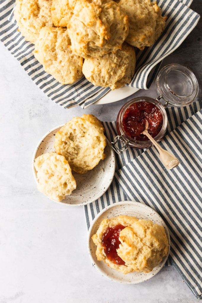 A plate of drop biscuits and several plates with biscuits being spread with butter and strawberry jam.