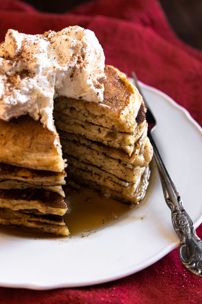 A stack of eggnog pancakes, topped with whipped cream and nutmeg, with a bite cut out.