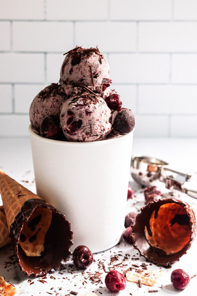 A container of black forest ice cream, with cones, cherries, and chocolate bits around.