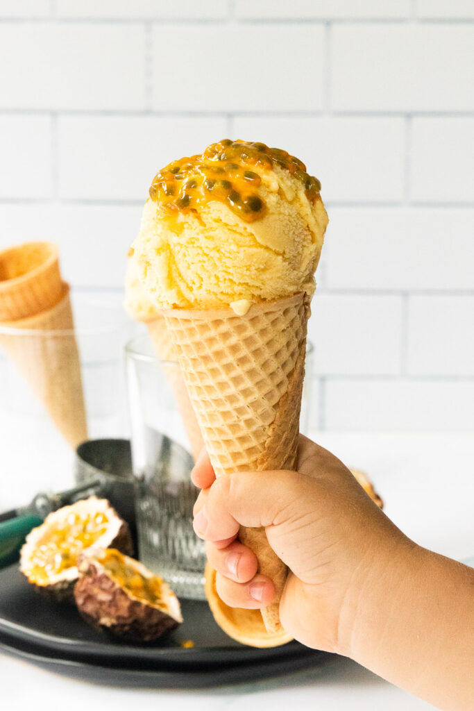 A scoop of passion fruit ice cream in a sugar cone, being held by a toddler's hand.