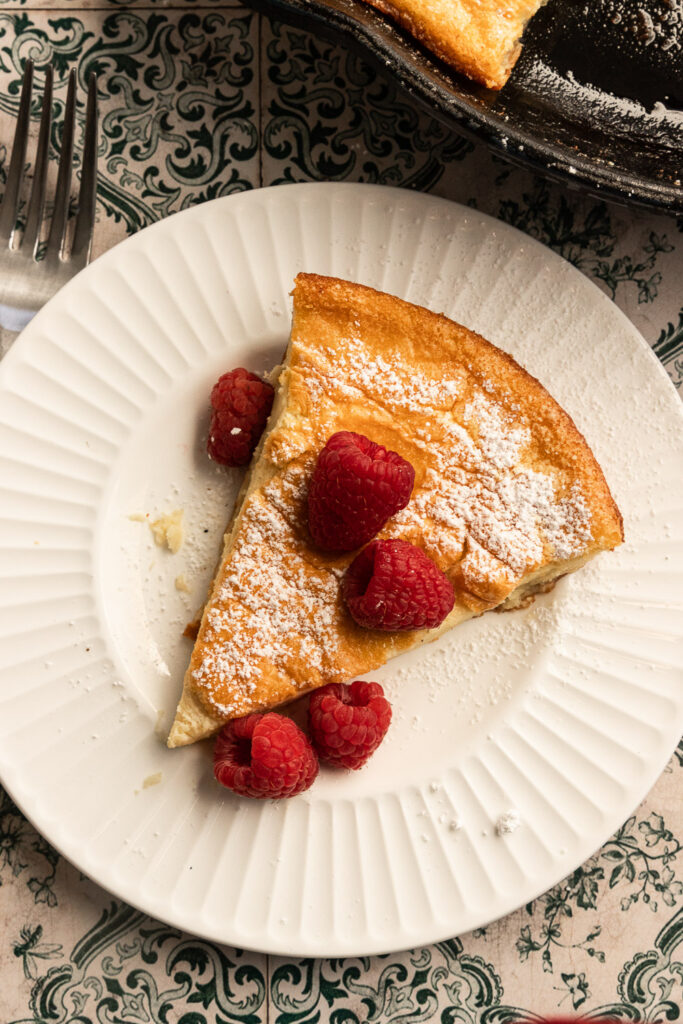 A slice of Sourdough Dutch Baby on a plate, topped with raspberries and powdered sugar.