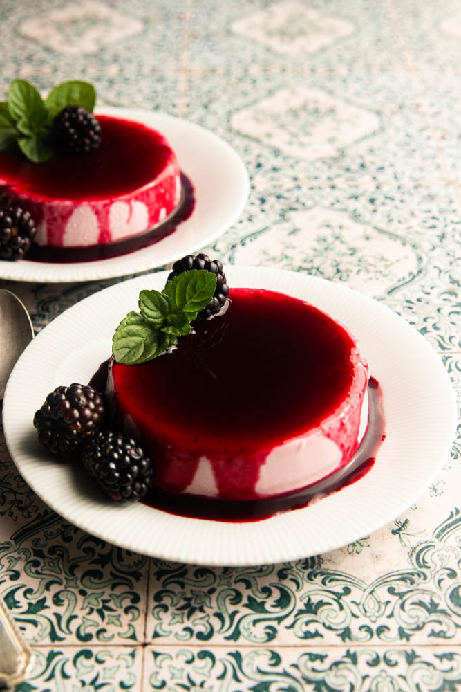 Blackberry panna cotta on a plate, covered with blackberry coulis and garnished with a fresh blackberry and mint leaves.