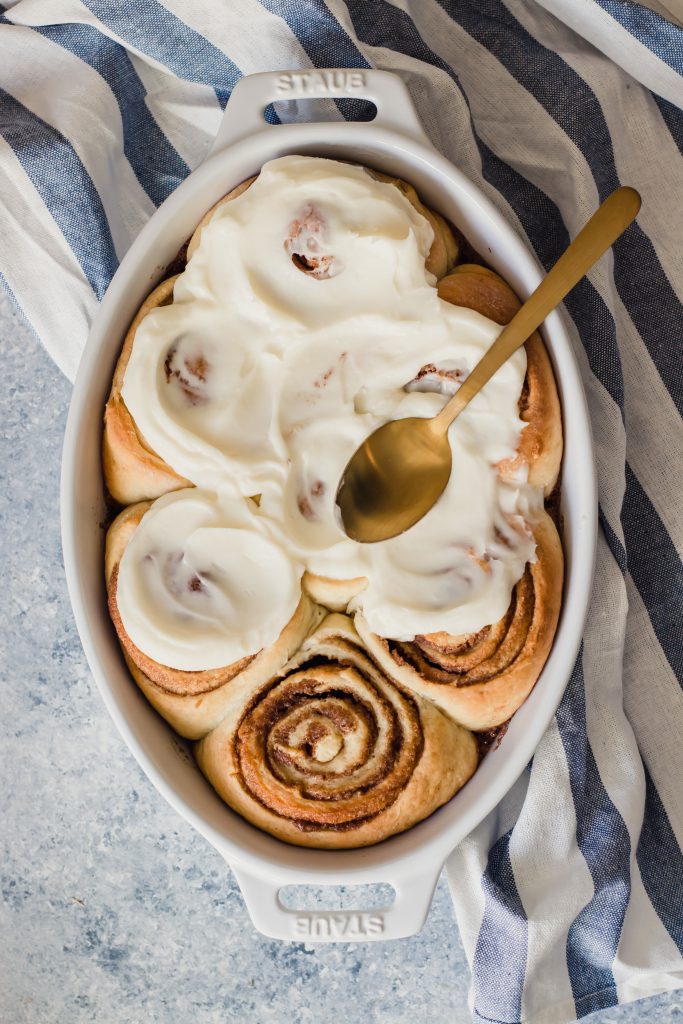 An oval pan of fresh, perfectly swirled cinnamon rolls with frosting being spread on them