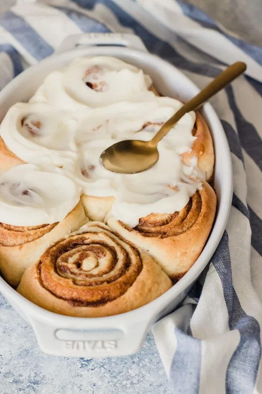 An oval pan of perfectly swirled cinnamon rolls being covered in fluffy white frosting.