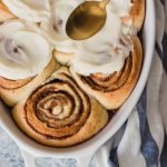 A pan of perfectly swirled cinnamon rolls with fluffy white frosting being spread over them.