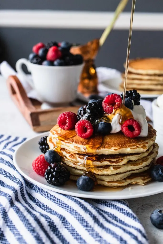 A stack of overnight sourdough pancakes, topped with whipping cream and berries, then with a drizzle of maple syrup.