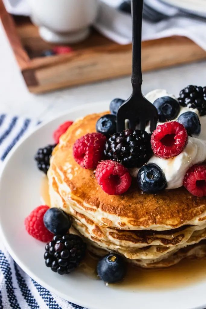 A stack of overnight sourdough pancakes, topped with whipping cream and berries, then with a drizzle of maple syrup.