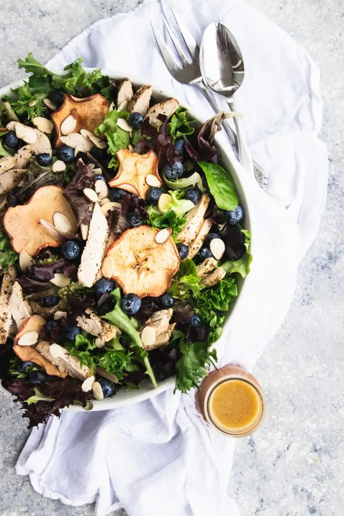 A giant bowl of fresh spring greens topped with dried apple chips, fresh blueberries, and slices of chicken. A small jar of fuji apple dressing is waiting to be pour over the salad.