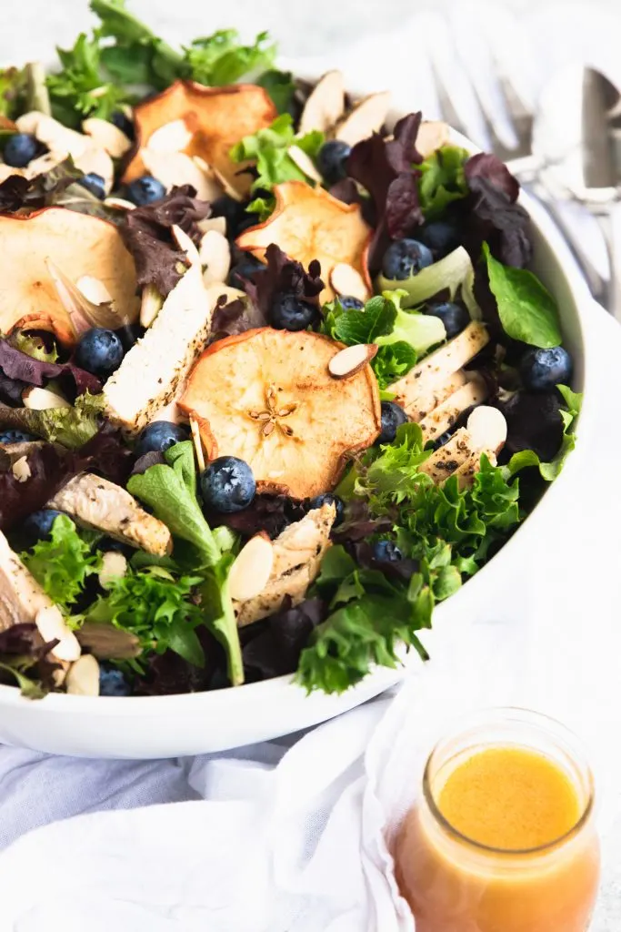 A giant bowl of fresh spring greens topped with dried apple chips, fresh blueberries, and slices of chicken. A small jar of fuji apple dressing is waiting to be poured over the salad.