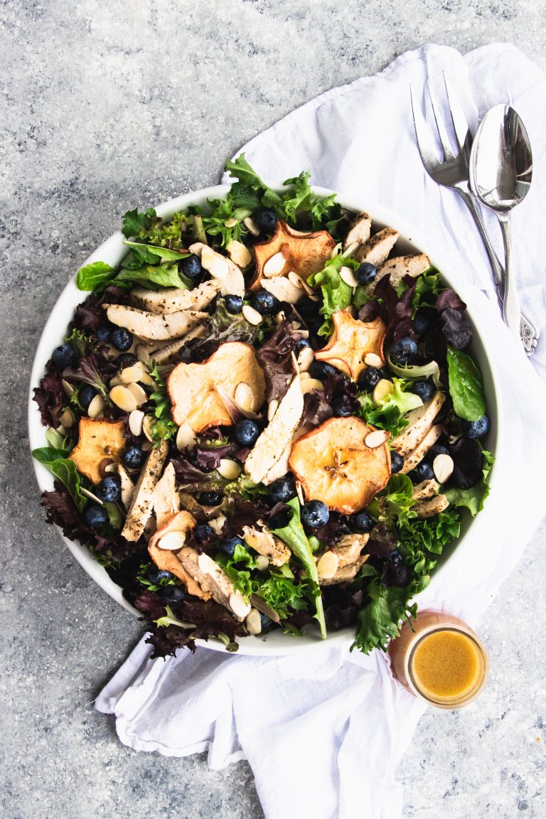 A giant bowl of fresh spring greens topped with dried apple chips, fresh blueberries, and slices of chicken. A small jar of fuji apple dressing is waiting to be poured over the salad.