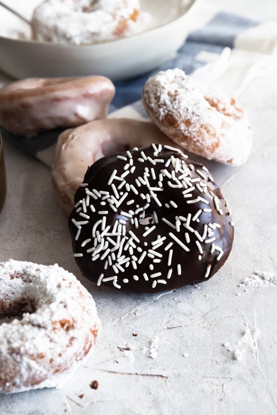 A pile of sourdough donuts covered in chocolate ganache and white sprinkles, powdered sugar, or glaze.