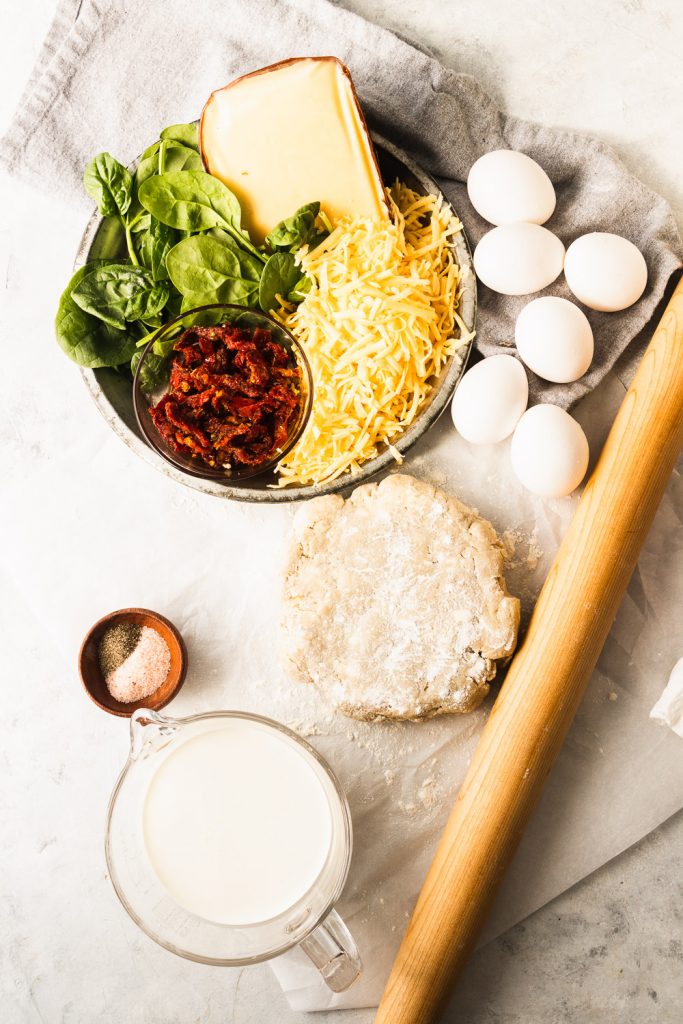 All of the ingredients for a sun dried tomato, spinach, and gouda quiche-- pie crust, eggs, milk, cream, shredded gouda, spinach, and sun dried tomatoes.