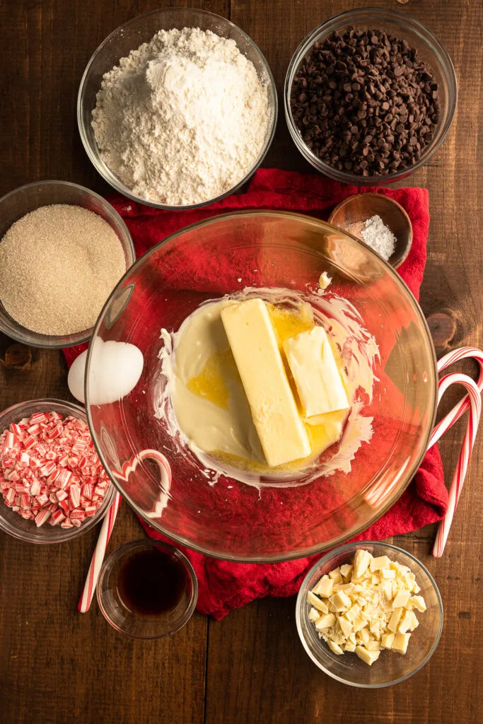 The ingredients for white chocolate peppermint cookies- butter, sugar, white chocolate, egg, vanilla, flour, baking powder, salt, candy canes, and mini chocolate chips.