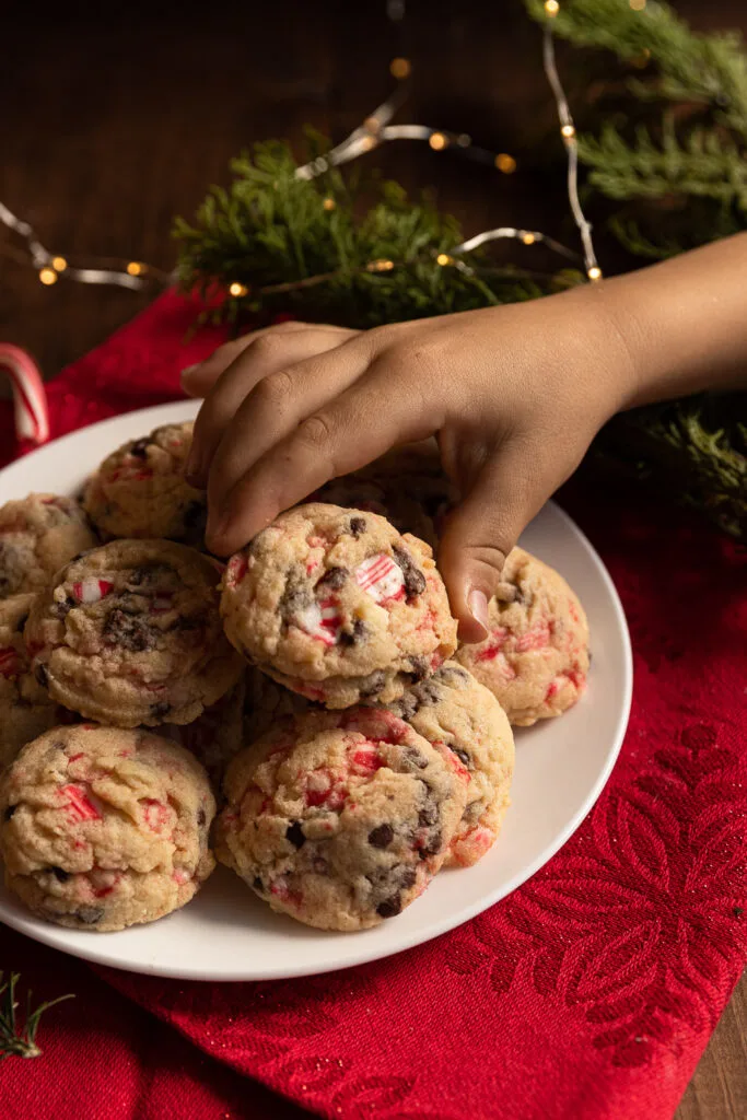 A child's hand picking up a white chocolate peppermint cookie from a plate.