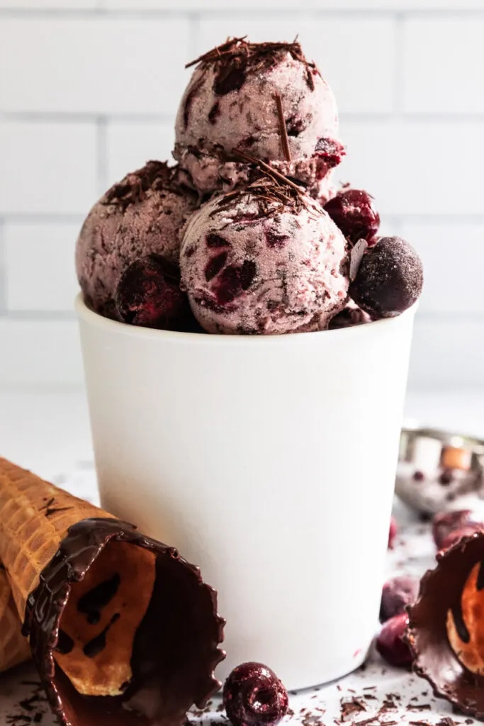 A container of black forest ice cream, with cones, cherries, and chocolate bits around.