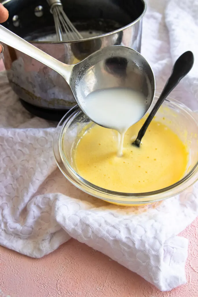 Whisking the hot milk into the egg yolks, a bit at a time, to warm them.
