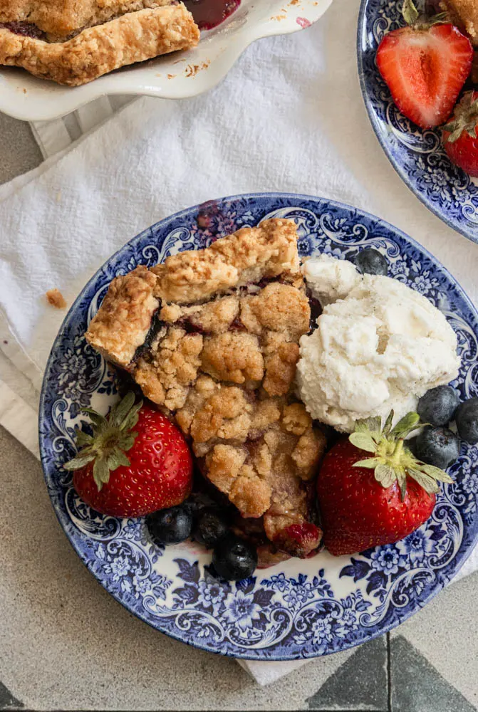 A piece of strawberry blueberry pie, served with fresh berries and a scoop of ice cream.